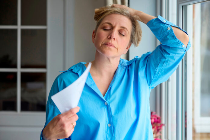 Menopausal Mature Woman Having Hot Flush At Home Cooling Herself With Letters Or Documents